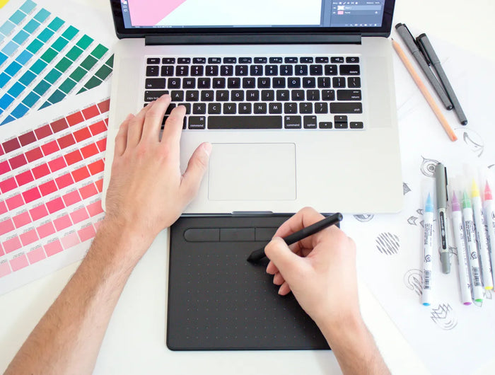 Person using a graphic tablet with a laptop on a desk, surrounded by color swatches and drawing tools.
